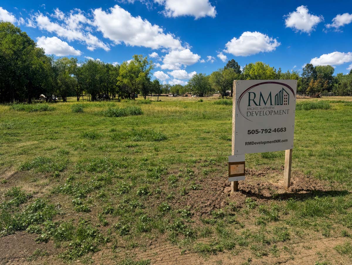 Open grassy vacant lot with a RM Development sign and a bright blue sky with white clouds in the background.